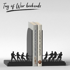Puede incluir: Sujetalibros de metal negro con dise&ntilde;o de tira y afloja. Los sujetalibros sostienen dos libros, "The Field Guide to Outdoor Games" y "A History of Team Sports." El texto "Tug of War bookends" est&aacute; escrito encima.