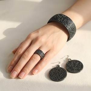 Puede incluir: Un conjunto de joyas a juego en gris oscuro, que incluye una pulsera, un anillo y pendientes. La pulsera es ancha, el anillo es una banda y los pendientes son circulares. Cada pieza tiene un patrón de laberinto repetitivo.