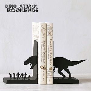 Puede incluir: Dos sujetalibros de metal negro con siluetas de dinosaurios. Una pieza muestra una cabeza de T-Rex frente a un grupo de personas corriendo, la otra muestra la cola del T-Rex. Sostienen dos libros titulados The Age of Dinosaurs y Paleontology Field Guide.