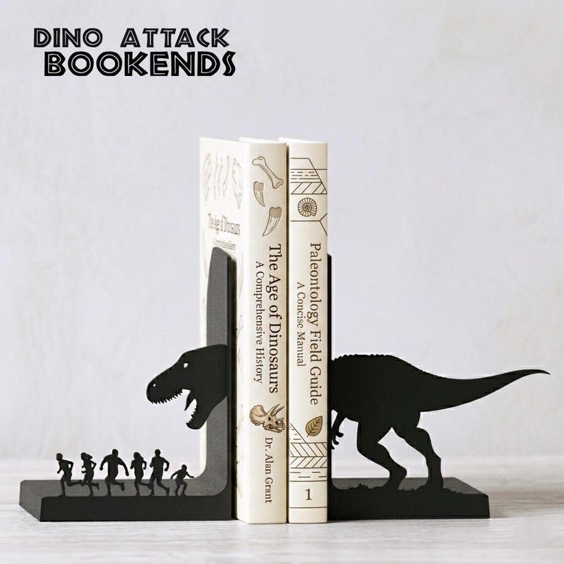 Puede incluir: Dos sujetalibros de metal negro con siluetas de dinosaurios. Una pieza muestra una cabeza de T-Rex frente a un grupo de personas corriendo, la otra muestra la cola del T-Rex. Sostienen dos libros titulados The Age of Dinosaurs y Paleontology Field Guide.