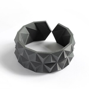 May include: A dark gray geometric bracelet with a repeating triangular pattern. The open cuff design features a series of raised, angular shapes, creating a textured surface. The bracelet is set against a white background.