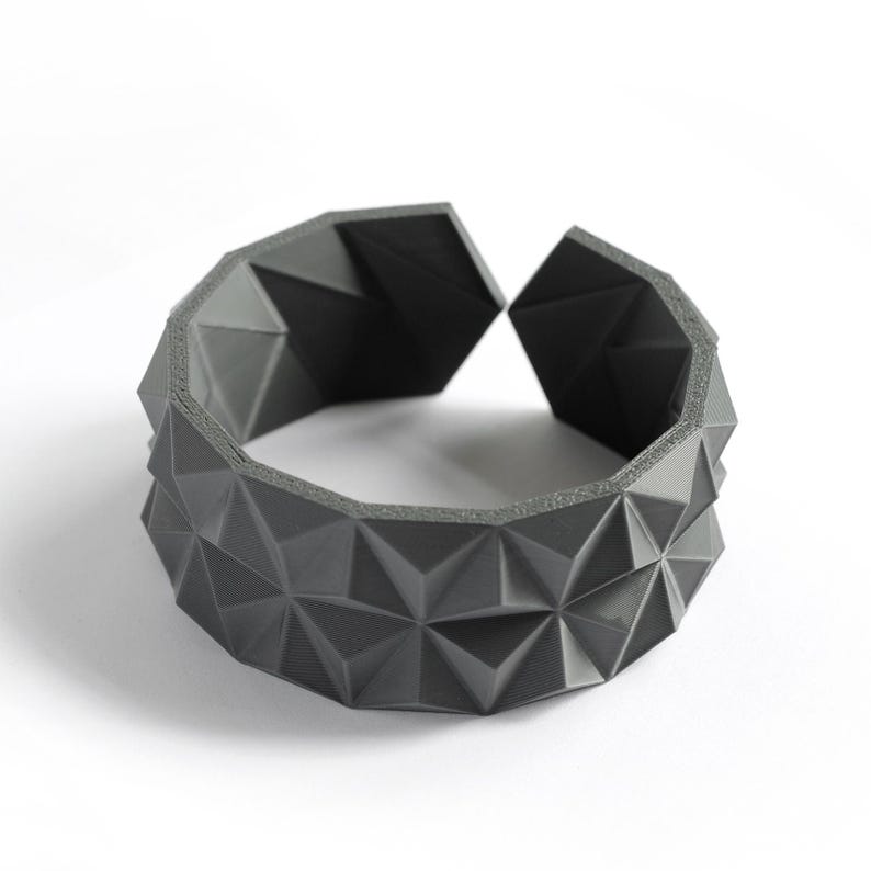 May include: A dark gray geometric bracelet with a repeating triangular pattern. The open cuff design features a series of raised, angular shapes, creating a textured surface. The bracelet is set against a white background.