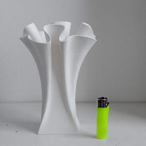 Puede incluir: Un jarr&oacute;n blanco mate con una forma alta, escultural y ondulada que presenta m&uacute;ltiples aberturas acampanadas en la parte superior. Junto al jarr&oacute;n hay un encendedor desechable de color verde ne&oacute;n brillante, que sirve de referencia.