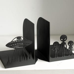 Puede incluir: Soportes para libros de metal negro. Uno presenta un dise&ntilde;o de OVNI con un alien&iacute;gena y una escena de bosque, el otro una familia de alien&iacute;genas. Dise&ntilde;ados para mantener los libros en posici&oacute;n vertical.