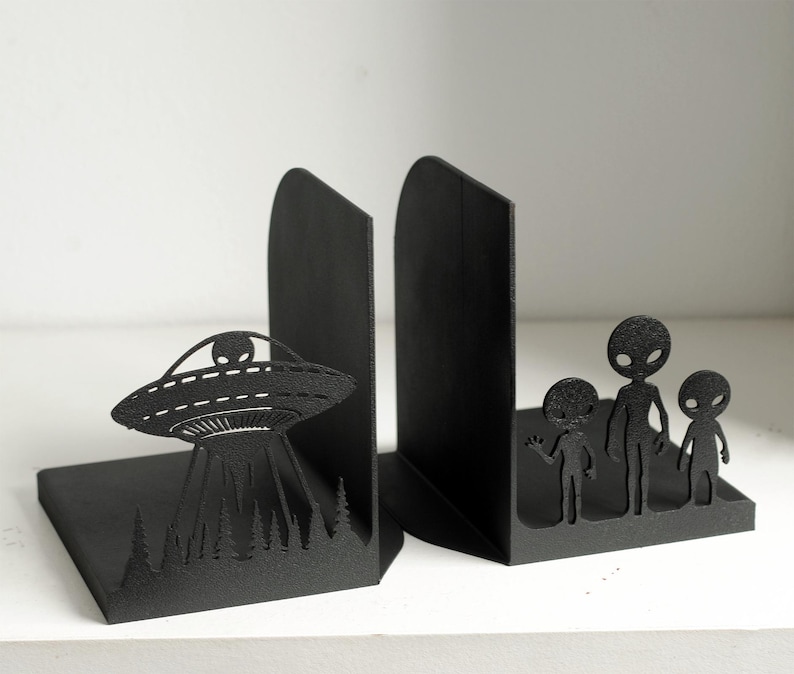 Puede incluir: Soportes para libros de metal negro. Uno presenta un dise&ntilde;o de OVNI con un alien&iacute;gena y una escena de bosque, el otro una familia de alien&iacute;genas. Dise&ntilde;ados para mantener los libros en posici&oacute;n vertical.