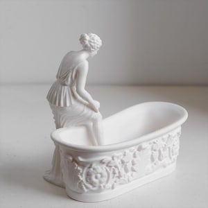 May include: A matte white ceramic figurine of a person with hair styled in an updo, seated on the edge of a small, oval-shaped bathtub. The exterior of the bathtub features a detailed relief pattern of floral motifs and scrolling foliage.