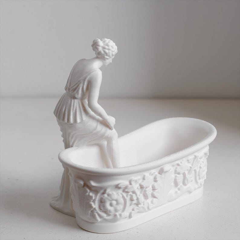 May include: A matte white ceramic figurine of a person with hair styled in an updo, seated on the edge of a small, oval-shaped bathtub. The exterior of the bathtub features a detailed relief pattern of floral motifs and scrolling foliage.