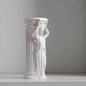 Puede incluir: Un florero cil&iacute;ndrico de color blanco mate que presenta una figura esculpida con vestiduras drapeadas de pie contra una columna estriada. La figura tiene ambos brazos levantados para sostener el borde superior, imitando un capitel cl&aacute;sico.