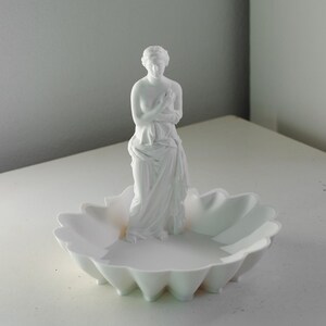 Puede incluir: Una bandeja decorativa de color blanco mate que presenta una figura central de pie envuelta en tela. La base de la bandeja tiene forma de flor festoneada con m&uacute;ltiples p&eacute;talos redondeados, proporcionando un cuenco poco profundo.