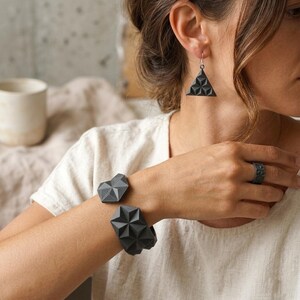 May include: A collection of geometric jewelry in dark gray. The set includes a triangular earring, a heart-shaped bracelet, a star-shaped bracelet, and a ring. The jewelry has a textured, three-dimensional design.