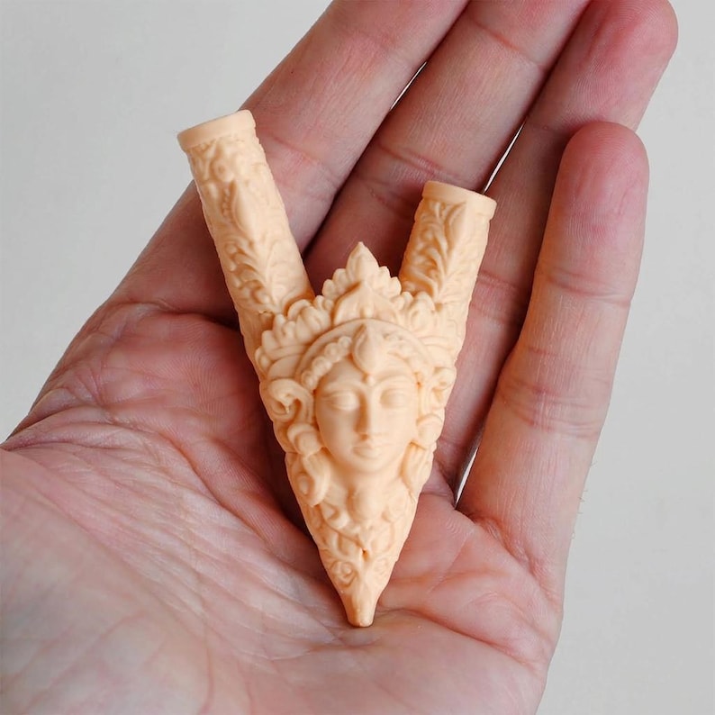 Puede incluir: Un objeto escult&oacute;rico en forma de V de color melocot&oacute;n claro, que presenta un relieve central de un rostro humano adornado con un tocado ornamentado. Los dos brazos superiores de la pieza tienen una textura con intrincados patrones de hojas.