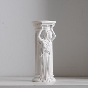 Puede incluir: Una escultura de pilar cil&iacute;ndrico de color blanco que presenta una figura esculpida con una prenda drapeada y los brazos levantados para sostener el capitel superior. La columna tiene estr&iacute;as verticales y un acabado mate y liso.