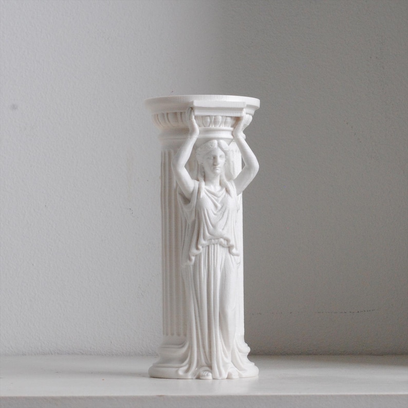 Puede incluir: Una escultura de pilar cil&iacute;ndrico de color blanco que presenta una figura esculpida con una prenda drapeada y los brazos levantados para sostener el capitel superior. La columna tiene estr&iacute;as verticales y un acabado mate y liso.