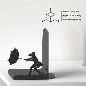 Puede incluir: Un sujetalibros de metal negro con la silueta de una persona sosteniendo un paraguas contra el viento. Las dimensiones son 11,5 cm de ancho, 12,6 cm de alto y 12,2 cm de profundidad. El dise&ntilde;o incluye una base plana y un panel de soporte.