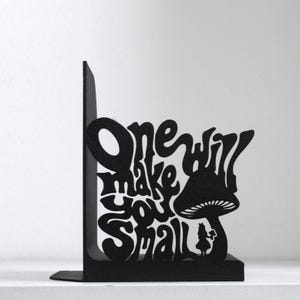 Puede incluir: Un sujetalibros de metal negro con un dise&ntilde;o recortado de una seta grande y una peque&ntilde;a figura de pie debajo. El texto "One will make you small" est&aacute; integrado en el dise&ntilde;o con una tipograf&iacute;a estilizada y fluida que ocupa el espacio.