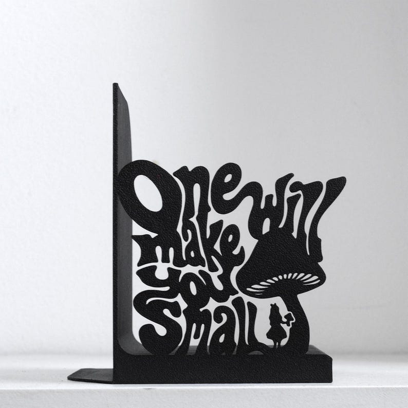 Puede incluir: Un sujetalibros de metal negro con un dise&ntilde;o recortado de una seta grande y una peque&ntilde;a figura de pie debajo. El texto "One will make you small" est&aacute; integrado en el dise&ntilde;o con una tipograf&iacute;a estilizada y fluida que ocupa el espacio.