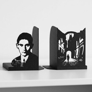 Puede incluir: Un par de sujetalibros de metal negro con dise&ntilde;os cortados con l&aacute;ser. Un sujetalibros muestra un retrato de Franz Kafka con su firma, mientras que el otro representa la silueta de una persona caminando bajo un arco de piedra.