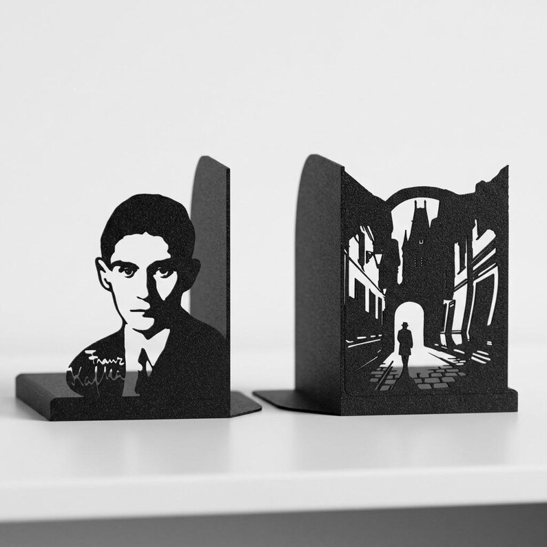 Puede incluir: Un par de sujetalibros de metal negro con dise&ntilde;os cortados con l&aacute;ser. Un sujetalibros muestra un retrato de Franz Kafka con su firma, mientras que el otro representa la silueta de una persona caminando bajo un arco de piedra.