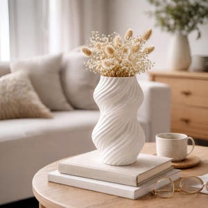Puede incluir: Un jarr&oacute;n de cer&aacute;mica blanca con un dise&ntilde;o retorcido, que contiene flores secas. El jarr&oacute;n est&aacute; colocado sobre una pila de libros en una mesa redonda de madera. Una taza y gafas tambi&eacute;n est&aacute;n sobre la mesa. El fondo incluye un sof&aacute; y un armario.