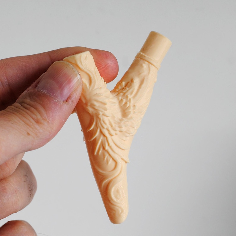 Puede incluir: Una pipa en forma de Y de color beige claro con una superficie texturizada que presenta intrincados relieves de plumas y patrones en espiral. El objeto tiene una abertura cil&iacute;ndrica en la parte superior y una base puntiaguda y c&oacute;nica.