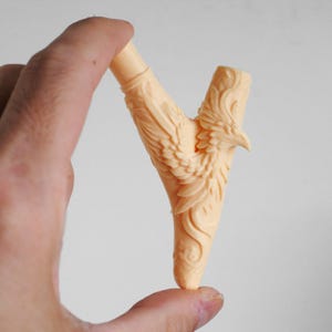 Puede incluir: Una pipa de fumar de color melocot&oacute;n claro que presenta una intrincada talla en relieve de un p&aacute;jaro con las alas extendidas. La pipa tiene una estructura en forma de Y con un tallo cil&iacute;ndrico en la parte superior y una cazoleta en el lado derecho, mostrando texturas de plumas detalladas y patrones.