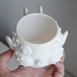 Puede incluir: Una maceta blanca impresa en 3D con forma de pez globo y centro hueco, que presenta una superficie texturizada con peque&ntilde;as p&uacute;as en relieve, dos ojos circulares prominentes y un peque&ntilde;o detalle de boca fruncida en el frente del cuerpo.