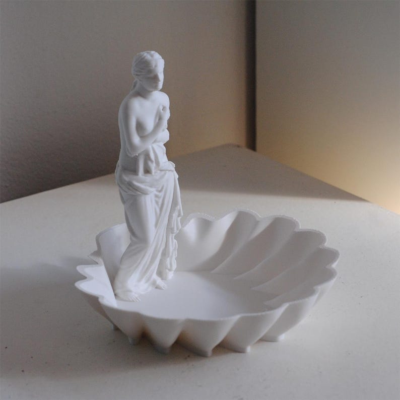 Puede incluir: Un cuenco decorativo de color blanco mate con forma de concha festoneada, que presenta una peque&ntilde;a figura cl&aacute;sica de pie de una persona drapeada unida a un lado. Todo el objeto es monocrom&aacute;tico, con un acabado liso y uniforme en la pieza.