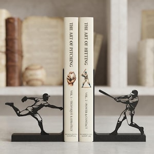 Puede incluir: Dos sujetalibros de metal negro con siluetas de jugadores de b&eacute;isbol, uno en posici&oacute;n de lanzamiento y otro en posici&oacute;n de bateo, sosteniendo dos libros titulados The Art of Pitching Vol. 1 y The Art of Hitting Vol. 2 con ilustraciones.