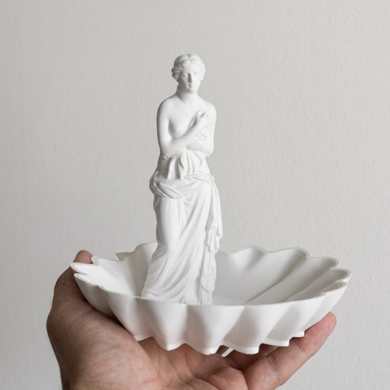Puede incluir: Una bandeja decorativa de color blanco mate que presenta una figura central de una estatua cl&aacute;sica de pie. La bandeja tiene un dise&ntilde;o de borde festoneado similar a una concha. Todo el objeto est&aacute; hecho de un material blanco s&oacute;lido y liso.
