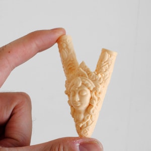 Puede incluir: Un objeto tallado en forma de V de color beige claro que presenta un relieve central de un rostro humano con patrones decorativos ornamentados y en espiral que se extienden por ambos brazos de la pieza. La superficie es lisa y mate.