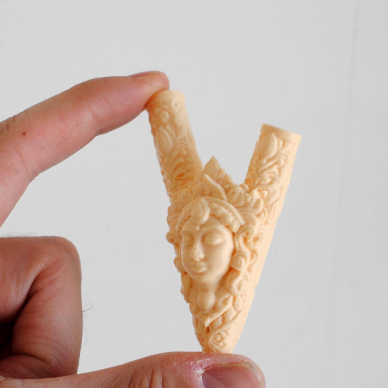 Puede incluir: Un objeto tallado en forma de V de color beige claro que presenta un relieve central de un rostro humano con patrones decorativos ornamentados y en espiral que se extienden por ambos brazos de la pieza. La superficie es lisa y mate.