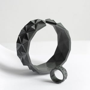 May include: A dark gray geometric bracelet and ring set. The bracelet is open and features a repeating triangular pattern. The matching ring is small and also has a geometric design. The jewelry is displayed on a white surface.