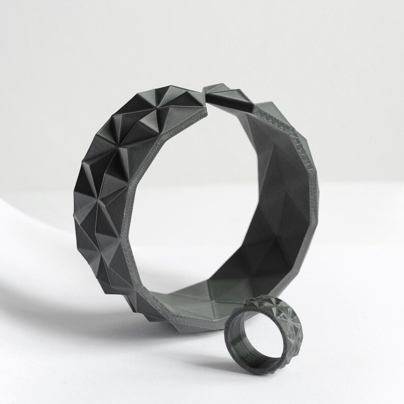 May include: A dark gray geometric bracelet and ring set. The bracelet is open and features a repeating triangular pattern. The matching ring is small and also has a geometric design. The jewelry is displayed on a white surface.