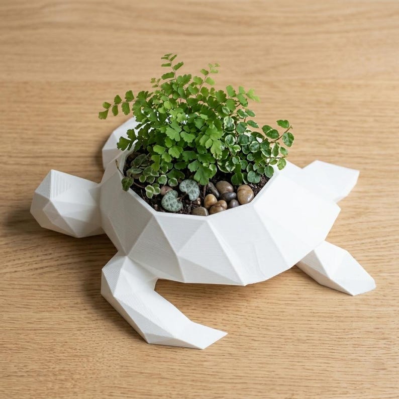 Puede incluir: Una maceta blanca con forma de tortuga geom&eacute;trica, llena de plantas verdes y peque&ntilde;os guijarros. La maceta tiene un dise&ntilde;o moderno y angular y est&aacute; colocada sobre una superficie de madera clara. La cabeza, las aletas y la cola de la tortuga est&aacute;n bien definidas.