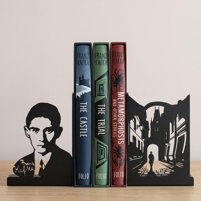 Puede incluir: Dos sujetalibros de metal negro con un retrato de Franz Kafka y la silueta de una persona caminando bajo un arco de piedra, flanqueando tres libros de tapa dura titulados The Castle, The Trial y Metamorphosis and Other Stories de Folio.