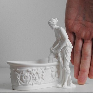 May include: A matte white ceramic figurine of a classical figure standing beside a bathtub. The bathtub features intricate relief carvings of floral and leaf motifs along its exterior side. The figure is depicted in a draped garment, lifting the fabric.