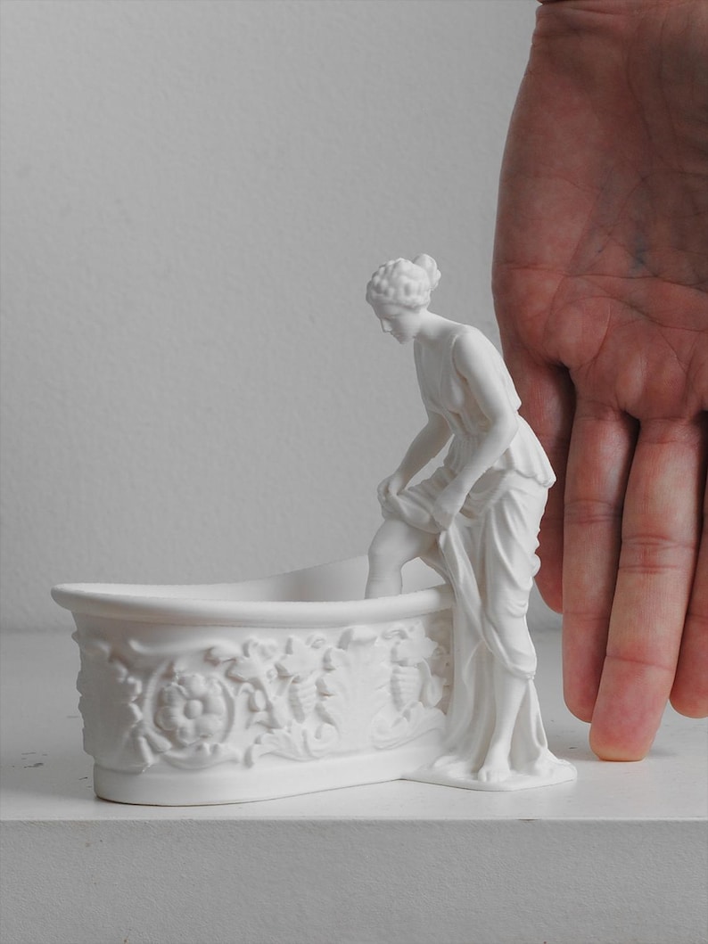 May include: A matte white ceramic figurine of a classical figure standing beside a bathtub. The bathtub features intricate relief carvings of floral and leaf motifs along its exterior side. The figure is depicted in a draped garment, lifting the fabric.