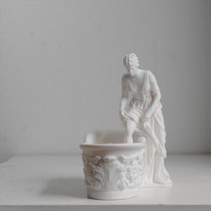 May include: A matte white ceramic figurine featuring a classical figure standing beside a small, ornate basin. The figure is depicted in a draped garment, with one leg raised as if stepping into the basin, which is decorated with relief carvings.