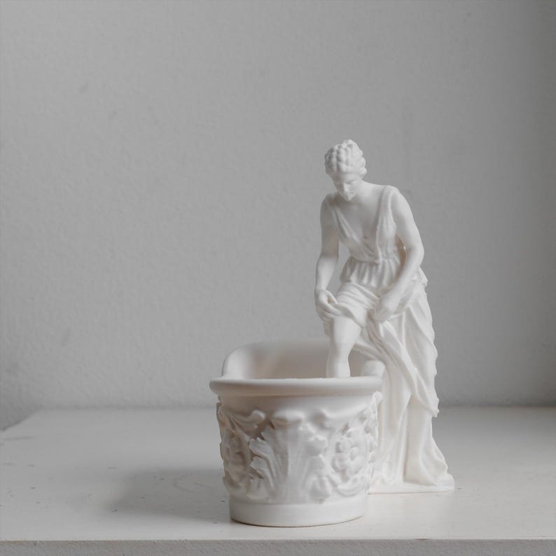 May include: A matte white ceramic figurine featuring a classical figure standing beside a small, ornate basin. The figure is depicted in a draped garment, with one leg raised as if stepping into the basin, which is decorated with relief carvings.