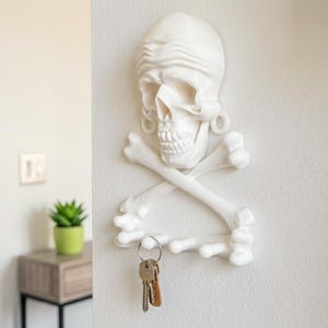 Puede incluir: Un soporte de pared blanco para llaves con forma de cr&aacute;neo humano, un pa&ntilde;uelo y huesos cruzados debajo. La estructura cuenta con cuatro ganchos salientes en la parte inferior para colgar objetos, con un llavero de metal colgado de uno de ellos.