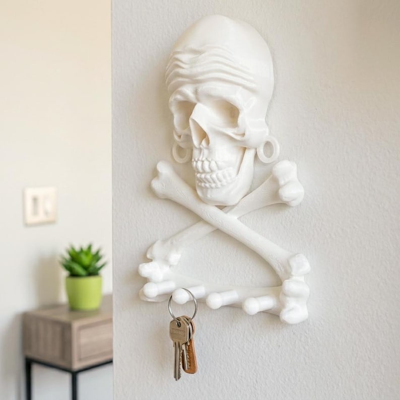 Puede incluir: Un soporte de pared blanco para llaves con forma de cr&aacute;neo humano, un pa&ntilde;uelo y huesos cruzados debajo. La estructura cuenta con cuatro ganchos salientes en la parte inferior para colgar objetos, con un llavero de metal colgado de uno de ellos.