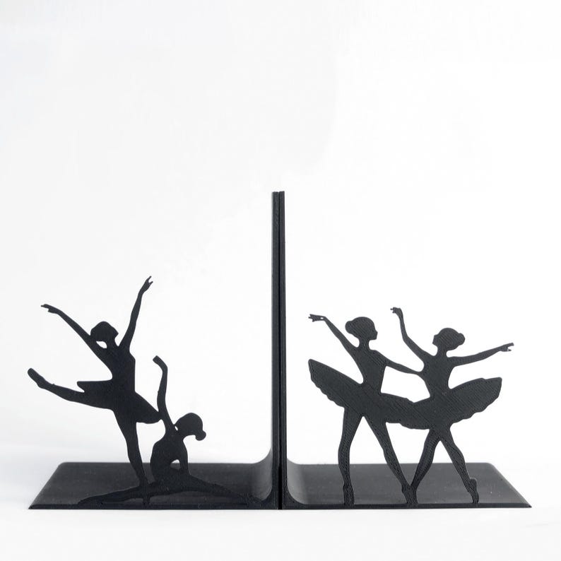 May include: Black metal bookends featuring silhouettes of ballerinas in various dance poses. The bookends have a rectangular base and a vertical support, with the dancers' forms cut out. The design is minimalist and elegant.