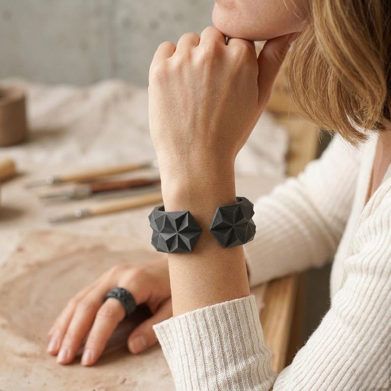 May include: Dark gray geometric cuff bracelet and ring set. The bracelet features two angular, starburst-like designs. The ring has a similar textured pattern. The jewelry is displayed on a person's wrist and finger.