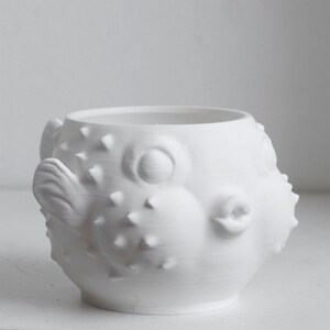 Puede incluir: Una taza de cer&aacute;mica blanca mate con forma de pez globo. El recipiente presenta un cuerpo redondeado con p&uacute;as puntiagudas en relieve, ojos circulares prominentes, una boca peque&ntilde;a fruncida y una aleta lateral. La superficie es lisa y uniforme.