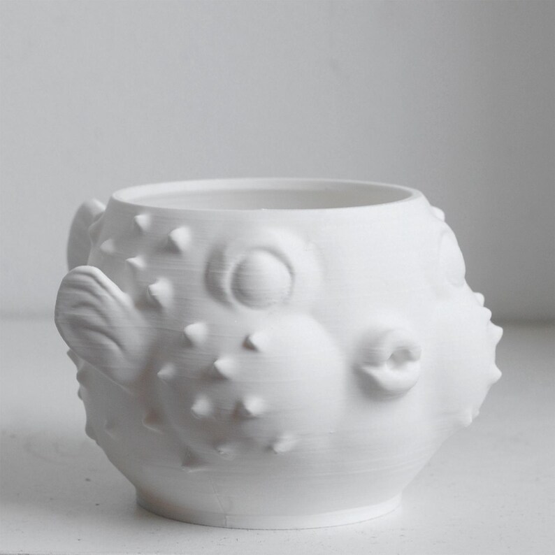 Puede incluir: Una taza de cer&aacute;mica blanca mate con forma de pez globo. El recipiente presenta un cuerpo redondeado con p&uacute;as puntiagudas en relieve, ojos circulares prominentes, una boca peque&ntilde;a fruncida y una aleta lateral. La superficie es lisa y uniforme.