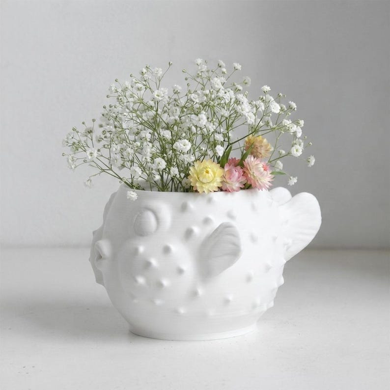 Puede incluir: Un jarr&oacute;n de cer&aacute;mica blanco mate con forma de pez globo que presenta una superficie texturizada y espinosa con un cuerpo redondeado. El jarr&oacute;n est&aacute; lleno de un ramo de peque&ntilde;as flores blancas de paniculata, junto con flores secas amarillas y rosas.