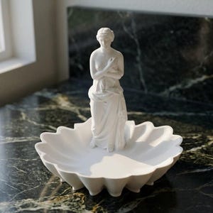 Puede incluir: Un plato decorativo de acabado blanco mate que presenta una figura central de pie con los brazos cruzados sobre el pecho. El plato tiene un dise&ntilde;o de borde festoneado similar a un p&eacute;talo y est&aacute; hecho de un material blanco s&oacute;lido, liso y opaco.