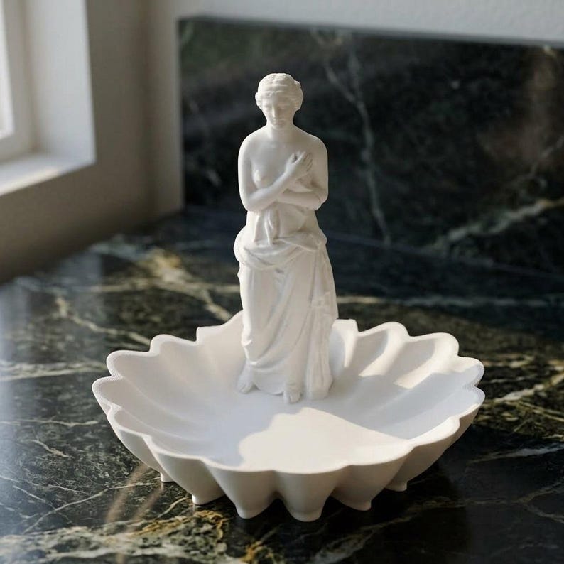 Puede incluir: Un plato decorativo de acabado blanco mate que presenta una figura central de pie con los brazos cruzados sobre el pecho. El plato tiene un dise&ntilde;o de borde festoneado similar a un p&eacute;talo y est&aacute; hecho de un material blanco s&oacute;lido, liso y opaco.