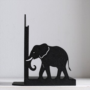 Puede incluir: Un sujetalibros de metal negro con la silueta de un elefante de perfil. El elefante est&aacute; colocado contra un panel de soporte vertical, con la trompa ligeramente curvada hacia arriba. El metal tiene un acabado mate con textura en toda la pieza.