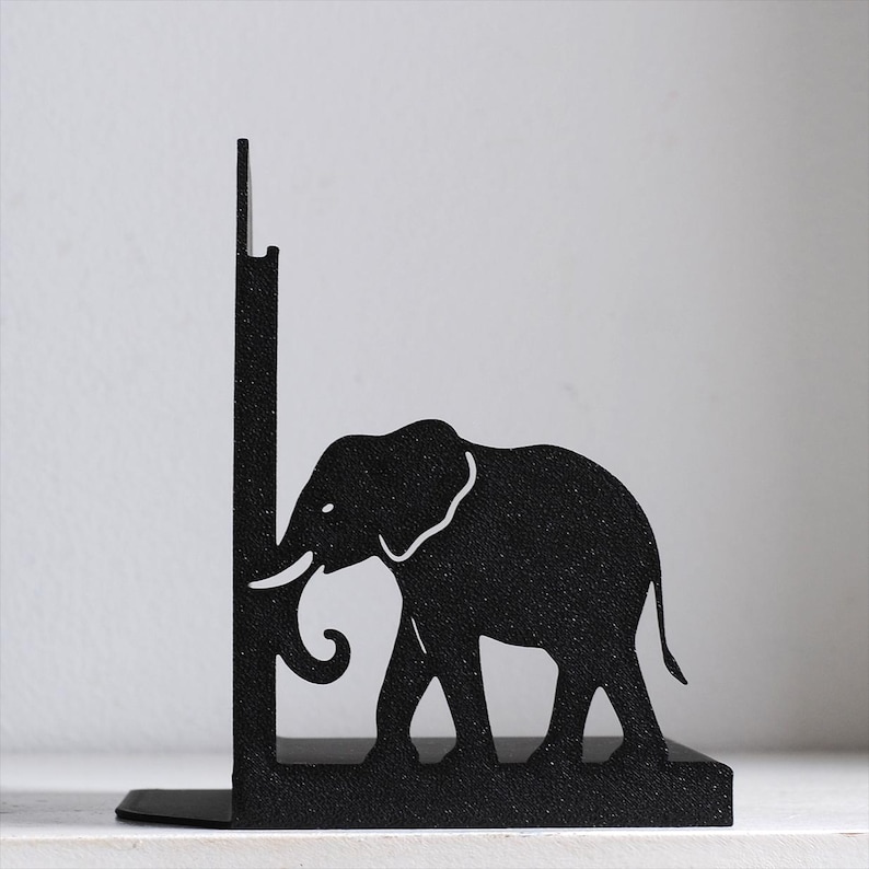 Puede incluir: Un sujetalibros de metal negro con la silueta de un elefante de perfil. El elefante est&aacute; colocado contra un panel de soporte vertical, con la trompa ligeramente curvada hacia arriba. El metal tiene un acabado mate con textura en toda la pieza.